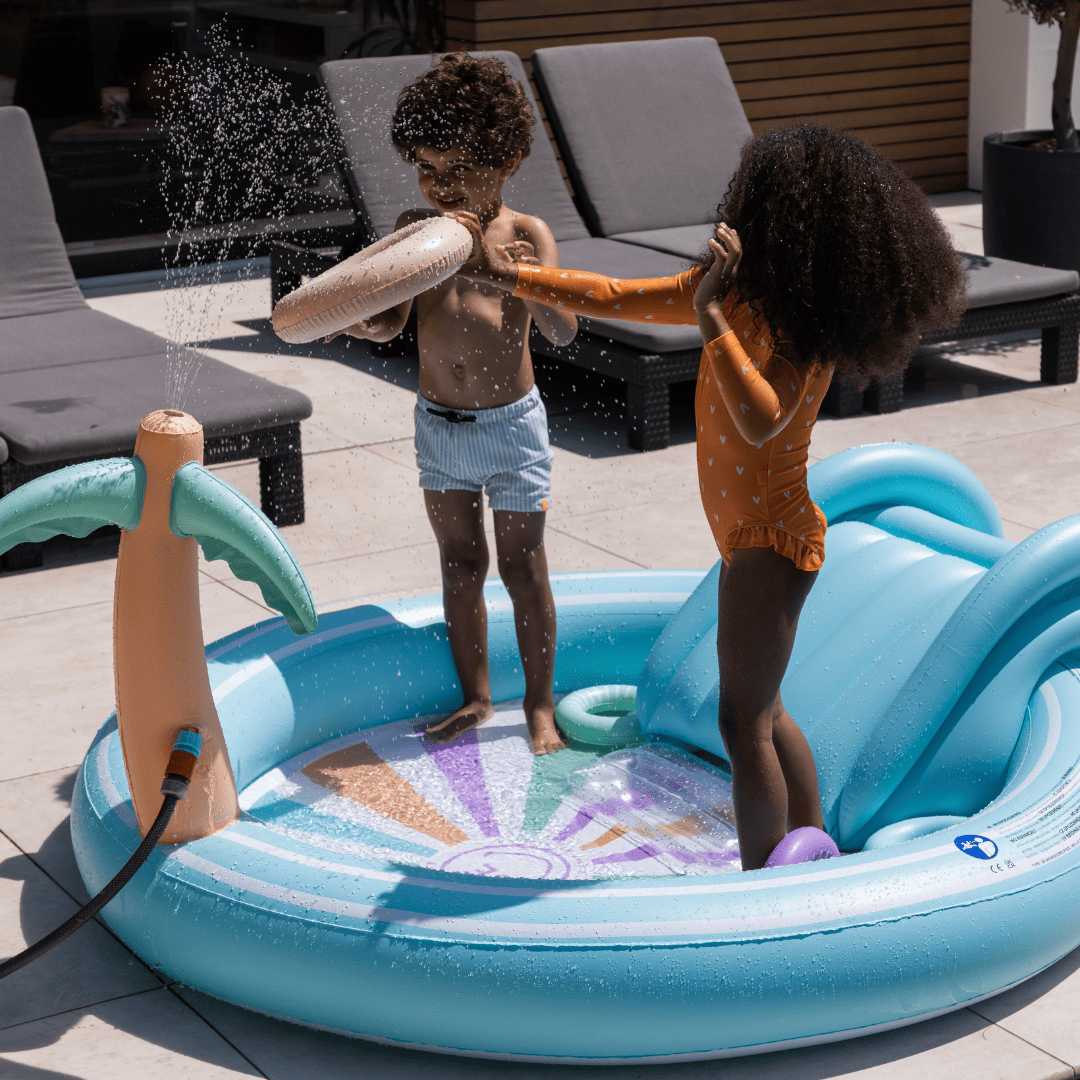 Two children playing in a blue inflatable pool with a water sprayer on a patio.