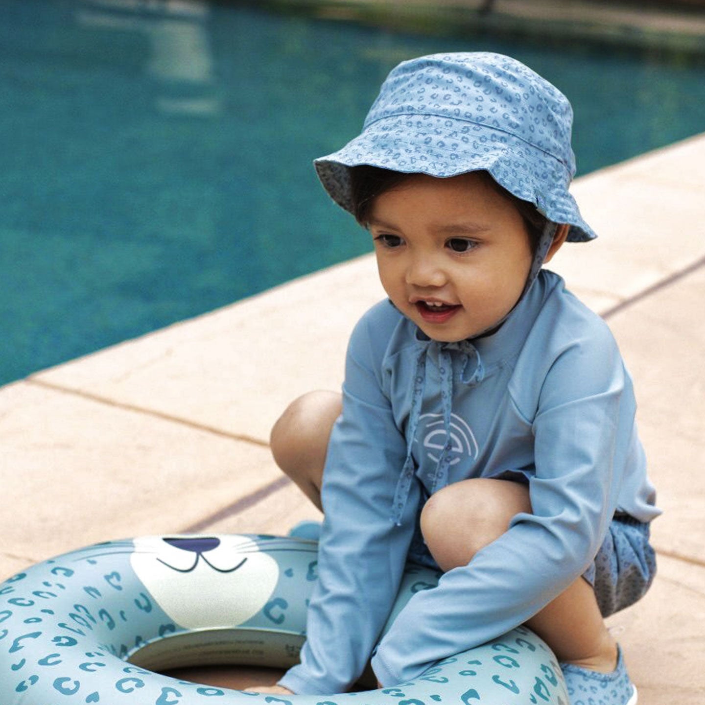 Child wearing a blue sun hat and swimsuit sitting on a pool float by a pool.