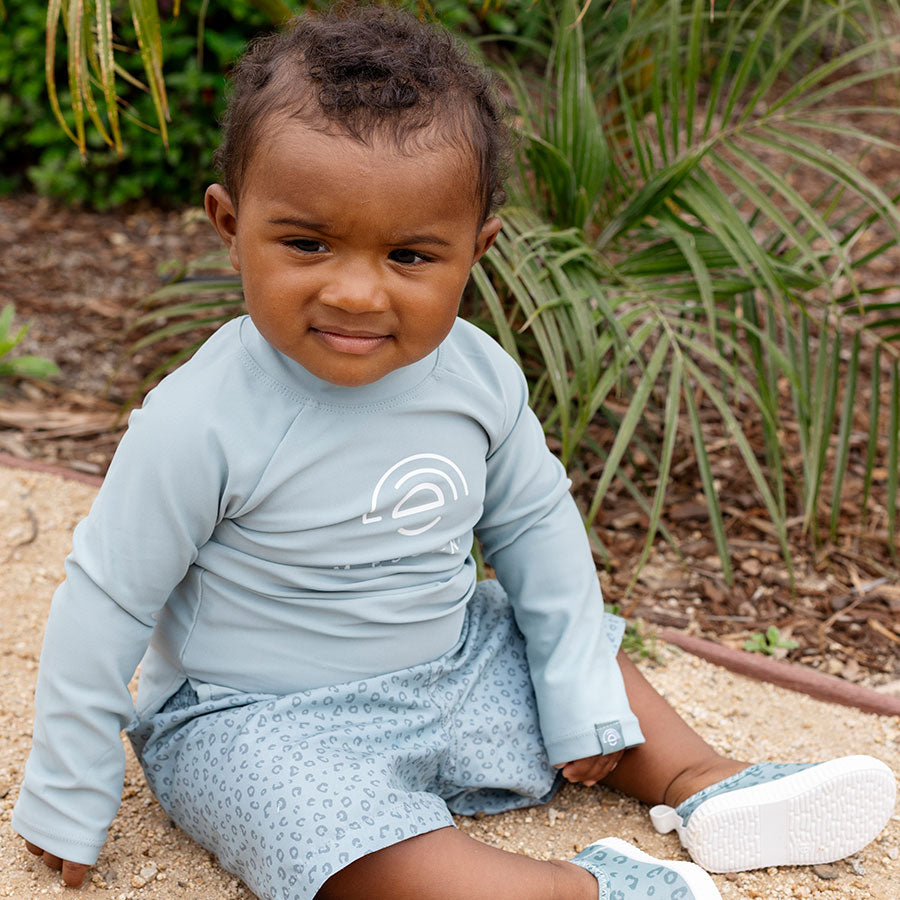 Child wearing a light blue long-sleeve shirt and shorts sitting outdoors with plants in the background