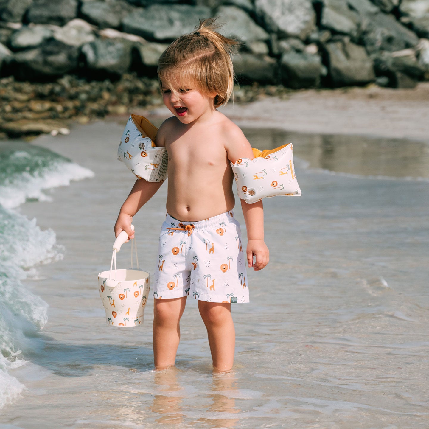 Child with arm floats and a bucket standing in shallow water at the beach.